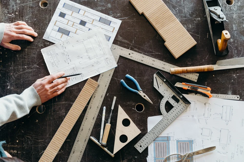 Workbench with measuring tools, hand plane, square, chisels, and a set of drawings and plans.